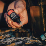 A hand squeezes Lifesystems Chlorine Dioxide Water Purification Droplets (2 x 30ml) from a bottle marked “2” over a black container, while another bottle labeled “1” by lifesystems sits on a patterned surface nearby.