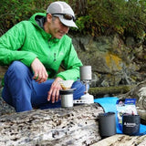 A person in a green jacket brews coffee with a camping stove and pour-over setup on a log, surrounded by outdoor gear—highlighting the versatility of the Soto Navigator Camping Cookware Set by Soto for outdoor adventures.