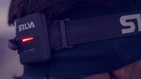 A close-up of a person wearing a headband with the Silva Trail Runner Free H Headlamp, featuring a small red LED display and supported by a hybrid battery.