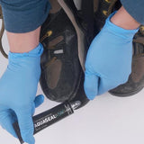 Hands in blue gloves expertly apply the Gear Aid Aquaseal Shoe Repair Glue from a tube to the sole of a worn hiking boot.
