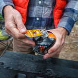 Hands opening a Jetboil Crunchit fuel Recycling Tool, wearing a flannel shirt and orange vest, with outdoor gear in the background.