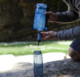 A person using a Sawyer Mini Water Filtration System to fill a clear bottle from a natural water source, amid clean water access in a wooded area.