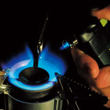 A metal spoon is heated over a blue gas flame, while a Soto Pocket Torch from Soto provides a wind-resistant, intensified flame aimed at the spoon.