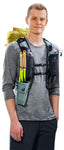 A person wears a gray long-sleeve shirt and black shorts, carrying the Deuter Vertrail 16 alpine pack by Deuter with a vest-style carry system, water bottles, and trekking poles stored in the front trekking pole holster.