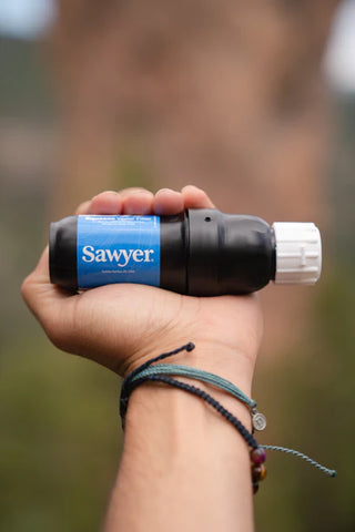 A hand holds the Sawyer Squeeze Water Filtration System by Sawyer outdoors with the label visible, highlighting this portable water filter against a blurred natural background.