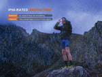 A person stands on a rock in the rain, using binoculars with a mountain backdrop. The Fenix Lighting HL12R V2.0 headlamp ensures visibility, while text highlights its IP66 waterproof and dustproof protection.