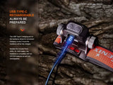 The Fenix Lighting HM65R-T V2.0 headlamp with Luminus SST40 & OSRAM LEDs is shown charging via USB Type-C, offering hands-free lighting and alternative battery compatibility—perfect for trail running needs.
