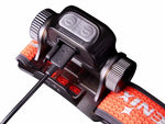 Close-up of the Fenix Lighting HM65R-T V2.0 headlamp (Luminus SST40 & OSRAM GW PUSTA1.PM LEDs) charging via USB, attached to its orange and black adjustable strap—ideal for hands-free lighting on any adventure.