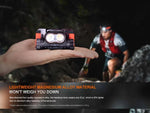A person holds the Fenix Lighting HM65R-T V2.0 headlamp with Luminus SST40 & OSRAM LEDs, while a runner in outdoor gear is blurred in the background. Text highlights its lightweight magnesium alloy for hands-free adventure lighting.