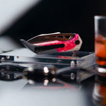 A Koi "Garry" Galah EDC Pocket Knife with a sleek red and black handle rests on a clear stand. To the right, a glass containing an orange beverage is partially visible, creating an ideal camping scene.