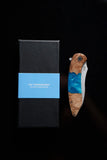 The Koi "Kyle" pocket knife by Koi Knives, featuring a wooden and blue resin handle, rests next to a black box marked with "The 'Kookaburra' Not Knives | South Australia" on a blue strip, ideal for outdoor explorers and EDC enthusiasts.