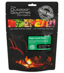 Packaging of the "Outdoor Gourmet Vegan Lentil Dahl" freeze-dried meal. The black bag from Outdoor Gourmet features text about the product, a green label indicating it's gluten-free and Halal/Kosher, along with images of food and a campfire scene.