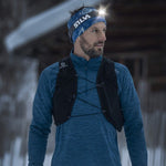 A man wearing a blue long-sleeve shirt and the Silva Trail Runner Free H Headlamp from the brand Silva, along with a blue headband, stands outdoors. With a black vest-like accessory on, he appears to be running in a snowy environment.