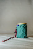 The Blue Tag Equipment Chalk Bag, featuring a small green design with a tan rim and red drawstring toggle, is displayed against a plain background.