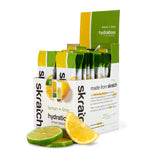 A Skratch Labs Sport Hydration Drink Mix Single Serving 20-Pack is shown with sliced lemon and lime in front. The open box displays single-serve electrolyte packets, ideal for convenient, on-the-go hydration during sports.