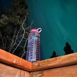 A Nalgene Nalgene Sustain Silo Bottle 1500ML sits on a wooden ledge with a backdrop of trees and an aurora-lit night sky, highlighting the beauty of sustainable water bottles made from recycled plastic bottles.