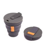 The Hunu Collapsible Cup by Hunu is shown in dark gray silicone with a lid and fabric band; a matching leak-proof lid with an orange tab lies beside this BPA-free cup.