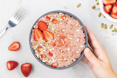 A hand holding a Radix Original 400kCal Breakfasts bowl of oatmeal topped with strawberries and seeds, perfect for athletes or anyone with an active lifestyle, with a fork and more strawberries scattered around on a marble surface.
