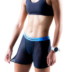 Model wears T8 Women's Commandos Running Underwear by T8, shown in black with a blue waistband and paired with a black sports bra, holding out the side of the shorts and accessorized with a white wristwatch.