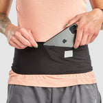 Wearing a peach shirt and gray shorts, a person slips their phone into the Ultimate Direction Utility Belt—a sleek black belt from Ultimate Direction that fits snugly around their waist without bouncing.
