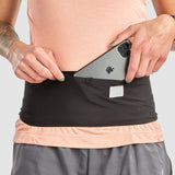 Wearing a peach shirt and gray shorts, a person slips their phone into the Ultimate Direction Utility Belt—a sleek black belt from Ultimate Direction that fits snugly around their waist without bouncing.