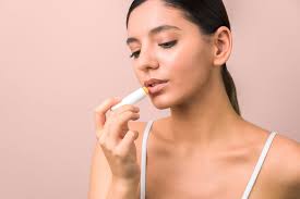 A woman applies Sunsational Lip Balm With SPF50+ Sunscreen by Sunsational to her lips against a neutral, light-colored background.