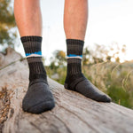 A person wearing dark gray Wilderness Wear Grampians Peak Merino eXtreme Hiker Socks with blue and white stripes stands on a fallen tree trunk outdoors.