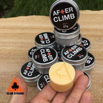 A hand holding a small container of Club Strong After Climb Hand Balm with Tea Tree oil, surrounded by other stacked containers, all displayed on grass.