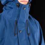 Close-up of a person wearing a Mont Men's Austral JKT by Mont, a blue hooded bushwalking rain jacket with drawstrings and black buttons. Made from Hydronaute fabric, it ensures waterproof breathable comfort. The face is partially obscured by the high collar and hood, against a black background.