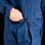 A person wearing a blue Mont Men's Austral JKT with their left hand in the jacket pocket. The jacket, made from Hydronaute fabric, features black velcro straps and zipper pulls visible, and a small Mont logo on the pocket.