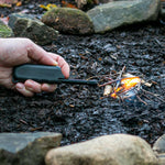 A hand using the Lifesystems Plasma Lighter, a windproof, rechargeable device by Lifesystems, to ignite a small pile of twigs on the ground. Stones are arranged around the fire.