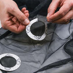 Hands applying a circular Gear Aid Tenacious Tape Mesh Patch 75mm to repair a tear on a black mesh fabric.